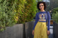 Woman wearing a blue top with a graphic design and a yellow skirt standing in front of green plants.