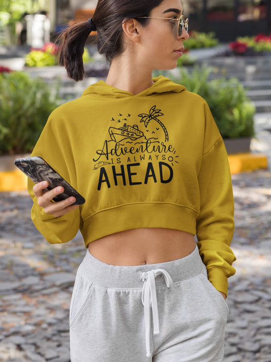 Woman wearing a mustard yellow hoodie with 'Adventure is Always Ahead' text, holding a phone, standing on a street.