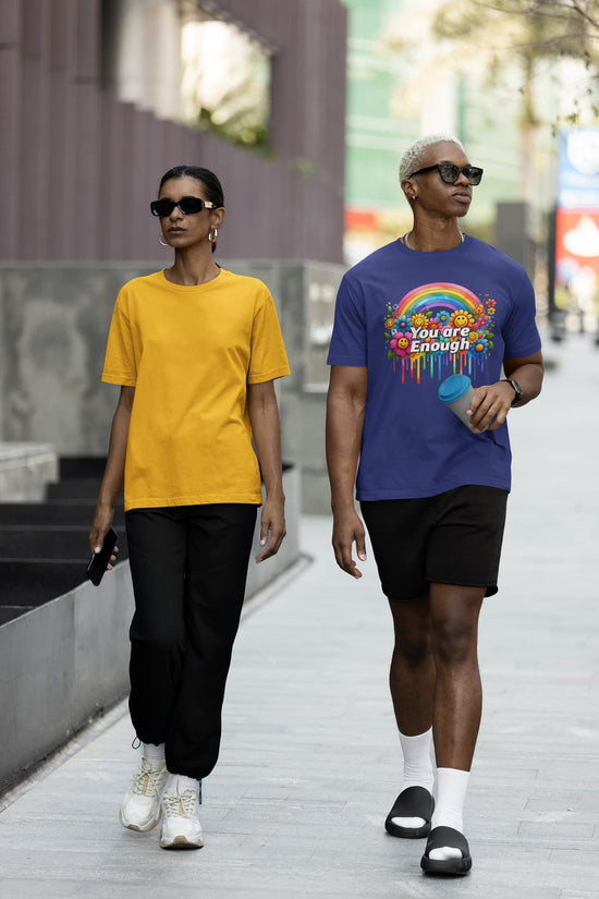 Two people walking on a sidewalk, one wearing a yellow shirt and black pants, the other wearing a blue shirt with a rainbow design and black shorts.