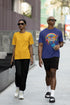 Two people walking on a sidewalk, one wearing a yellow shirt and black pants, the other wearing a blue shirt with a rainbow design and black shorts.