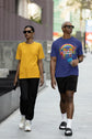 Two people walking on a sidewalk, one wearing a yellow shirt and black pants, the other wearing a blue shirt with a rainbow design and black shorts.
