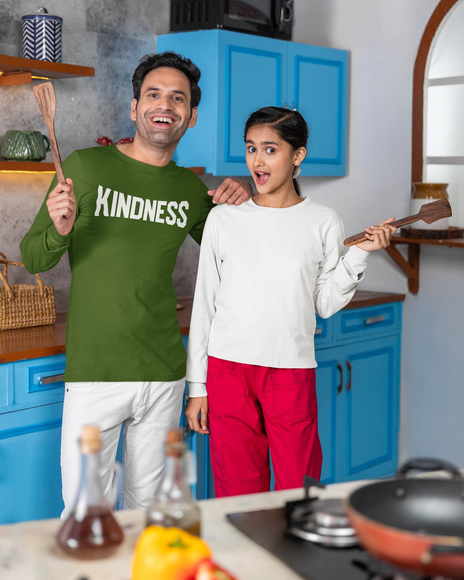 Man and woman in a kitchen holding spatulas, with the man wearing a green shirt with &
