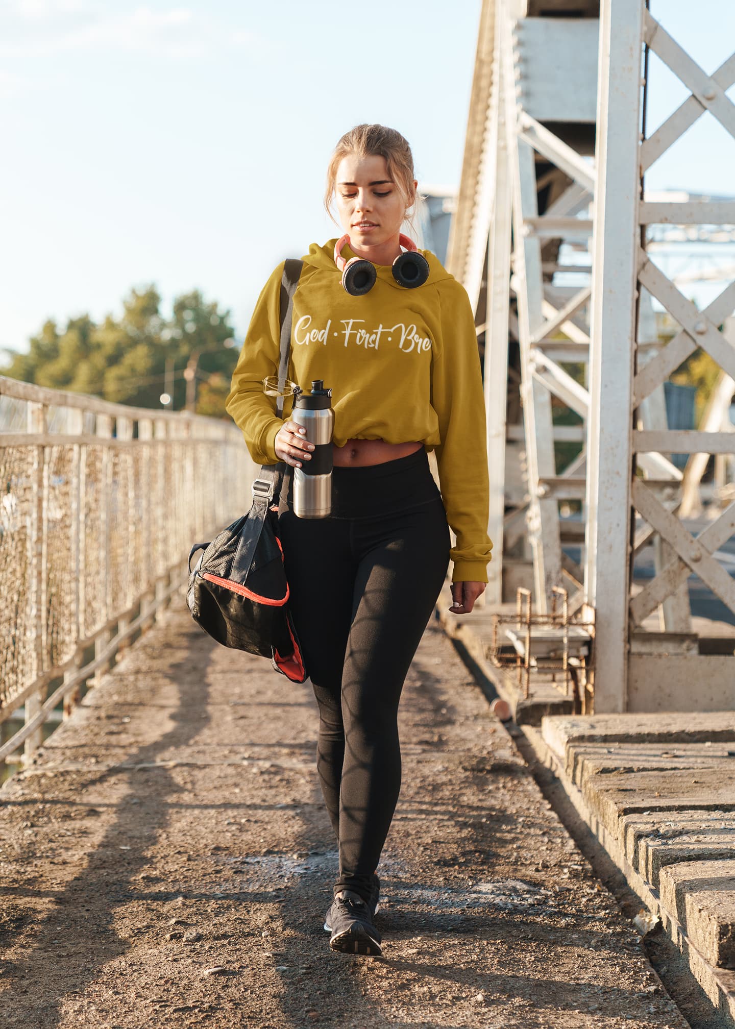 Woman walking outdoors carrying a water bottle and a bag, wearing a mustard yellow hoodie with text.