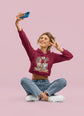 Woman taking a selfie wearing a maroon hoodie with a graphic design on a pink background