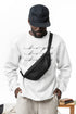 Man wearing a white sweatshirt with text, black cap, and black crossbody bag on a white background
