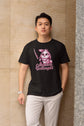 Person wearing a black t-shirt with a graphic design and text, standing against a textured wall.