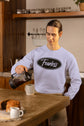 Person pouring coffee in a kitchen wearing a sweatshirt with &