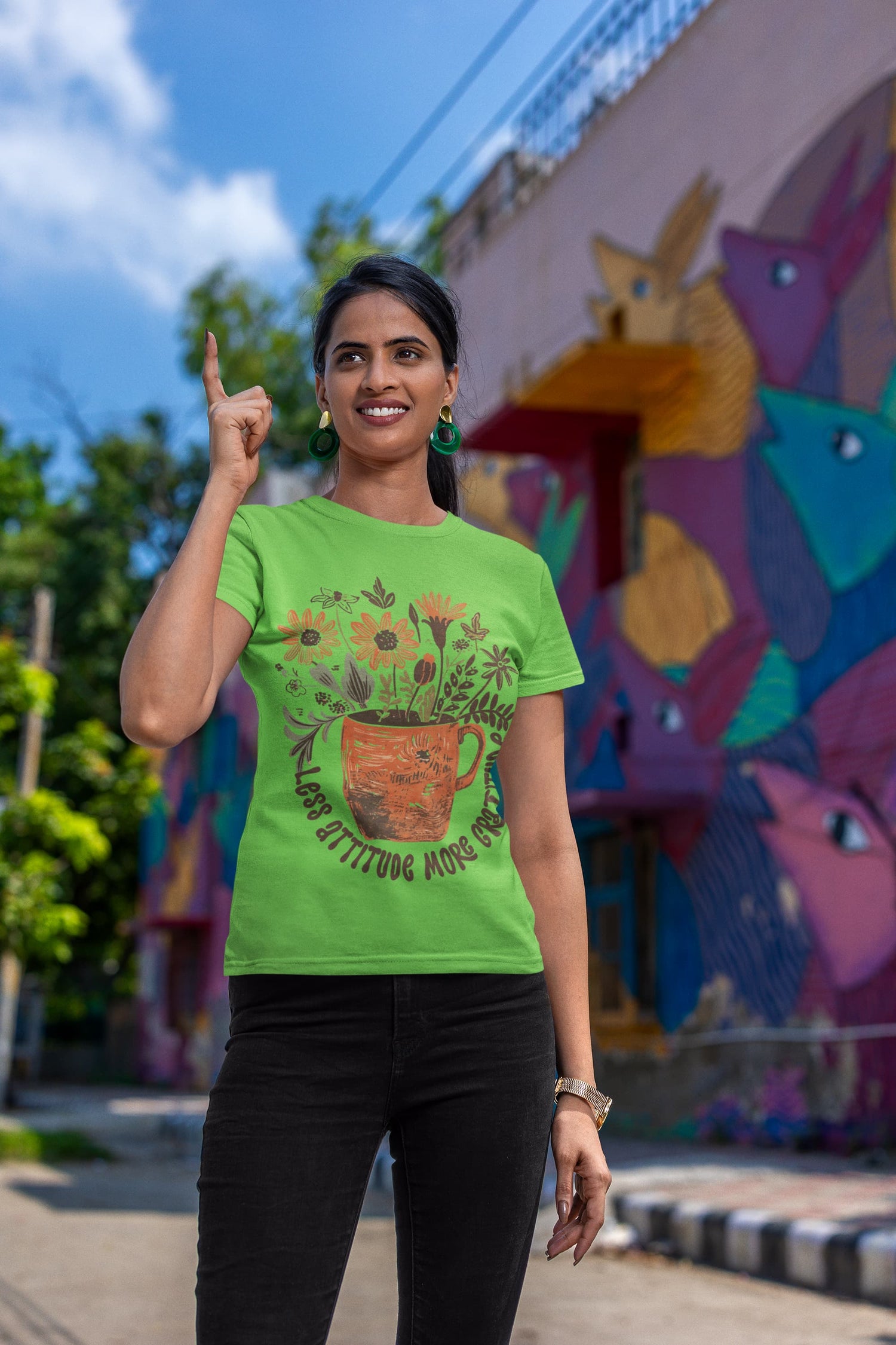 Woman wearing a green t-shirt with a floral design, pointing upwards in front of a colorful mural.