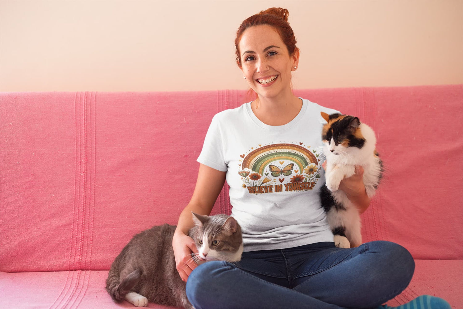 Woman wearing a t-shirt with a cat-themed design, sitting on a pink couch with two cats.