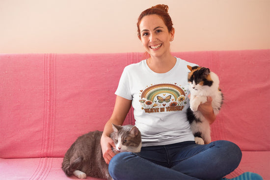 Woman wearing a t-shirt with a cat-themed design, sitting on a pink couch with two cats.