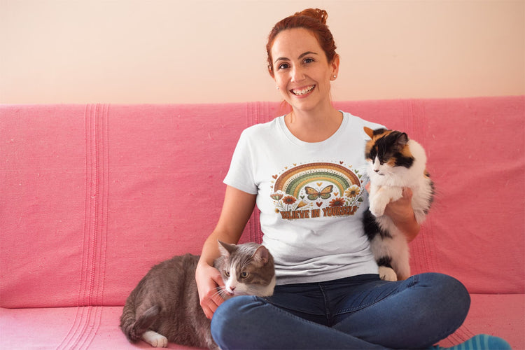 Woman wearing a t-shirt with a cat-themed design, sitting on a pink couch with two cats.