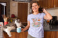 Woman in a kitchen wearing a t-shirt with a cat graphic and text, holding a mug.