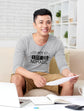 Man sitting on a couch with a laptop and papers, wearing a gray shirt with &
