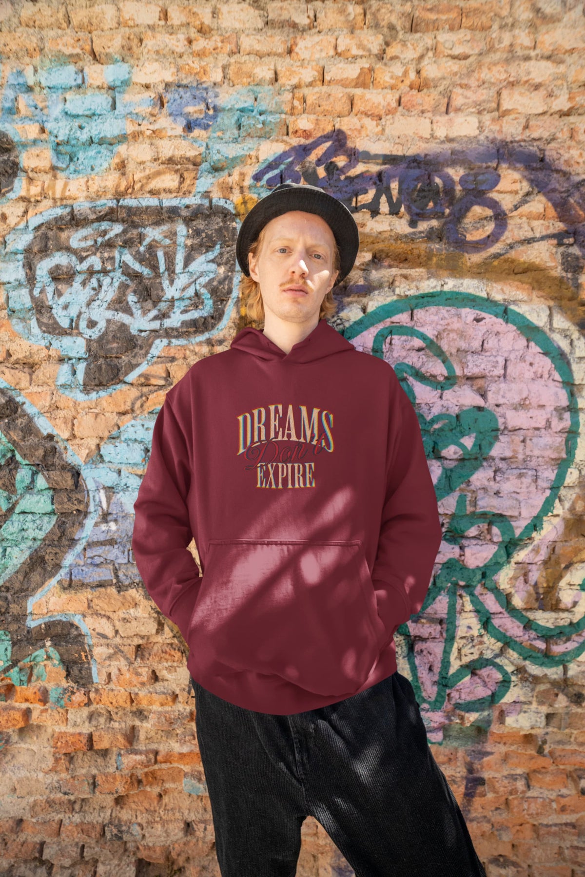 Person wearing a maroon hoodie with 'DREAMS EXPIRE' text against a graffiti-covered brick wall.