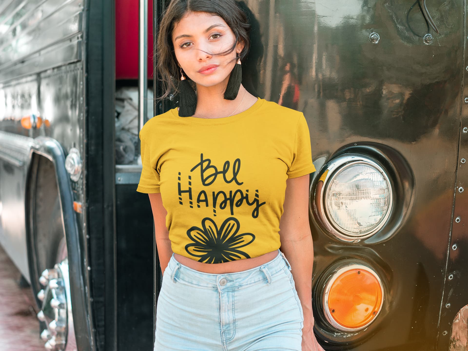 Woman wearing a yellow t-shirt with 'Be Happy' text standing in front of a vehicle.