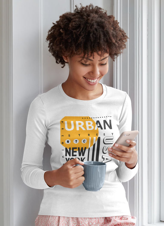 Woman wearing a white long-sleeve shirt with a graphic design, holding a mug and phone, standing against a light-colored wall.