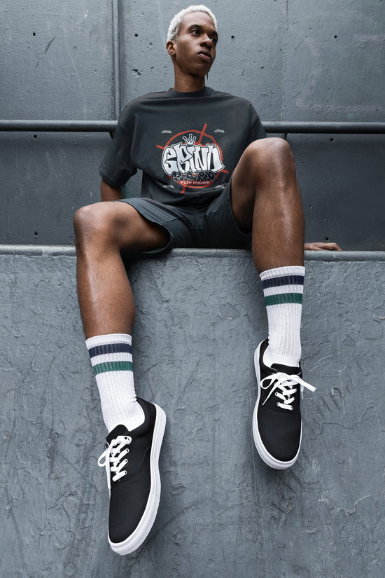 Person wearing black sneakers, white socks with stripes, and a dark t-shirt sitting against a gray wall.