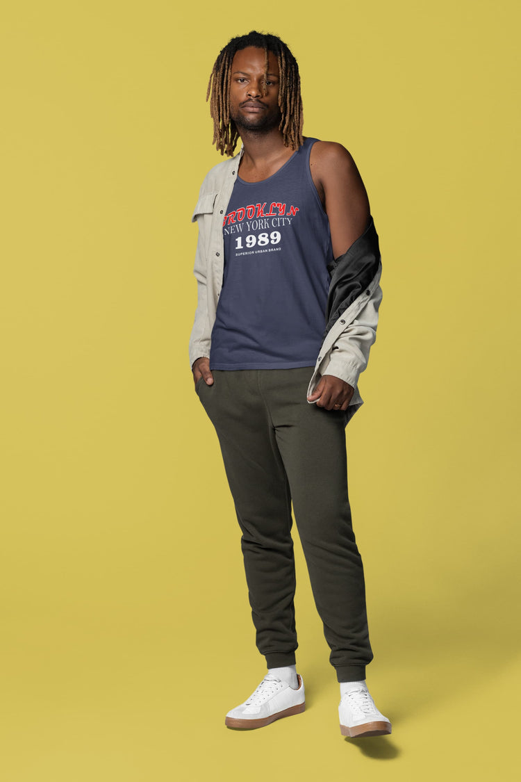 Man wearing a navy sleeveless shirt with text, beige jacket, green pants, and white sneakers on a yellow background