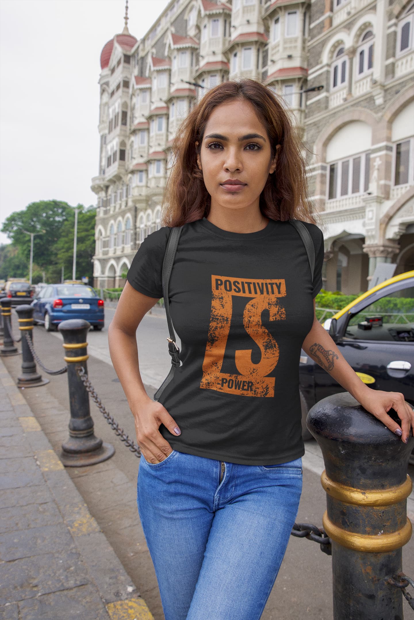 Woman wearing a black t-shirt with orange text and design, standing on a street with buildings in the background.