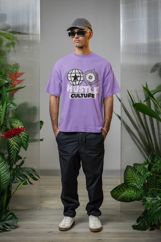 Person wearing a purple t-shirt with text and graphics, standing indoors with plants in the background