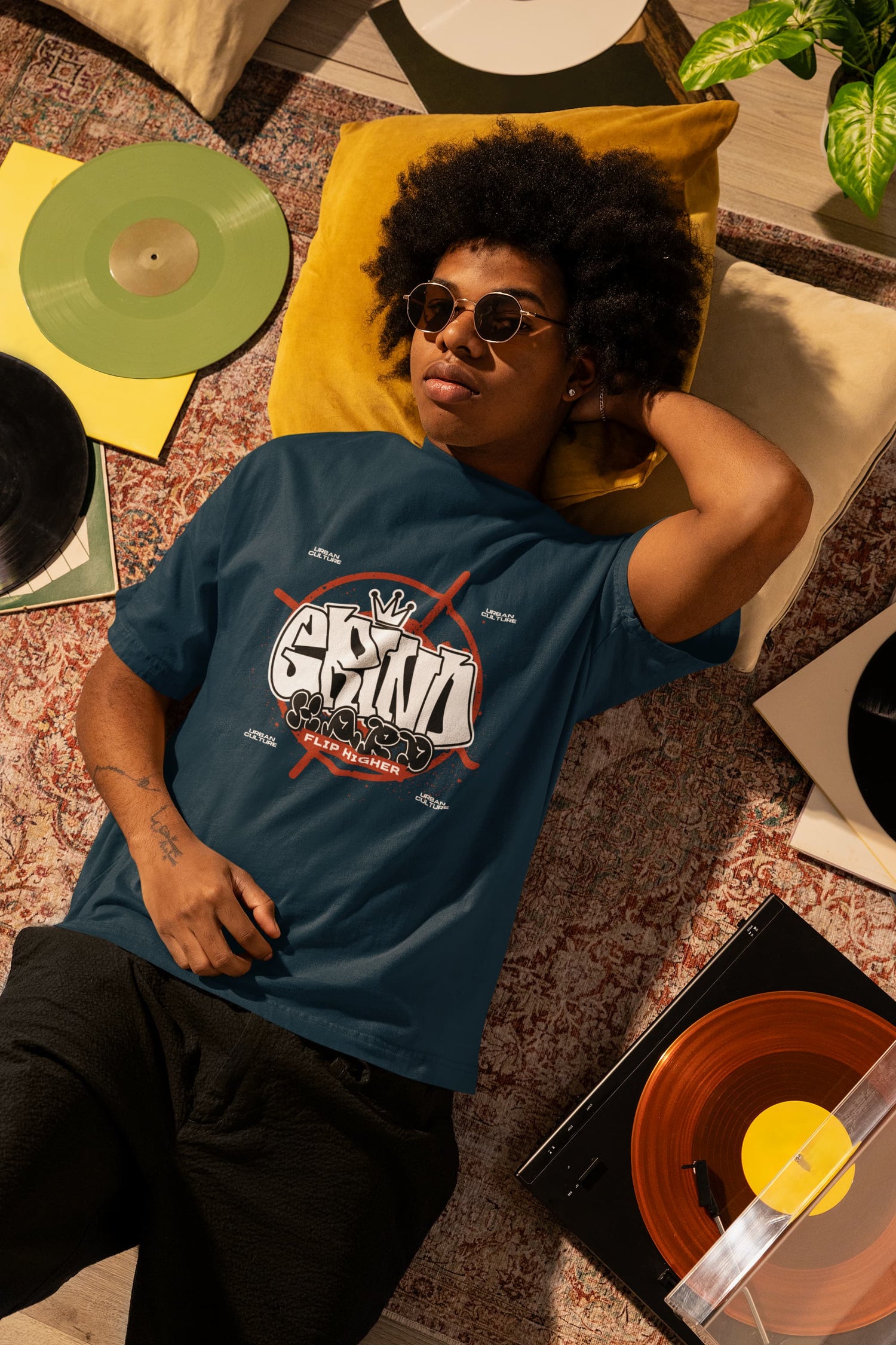 Person wearing a blue t-shirt with a graphic design, lying on a patterned floor with vinyl records and a plant.