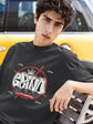 Person wearing a black t-shirt with graphic design, sitting on a bench with a yellow vehicle in the background.