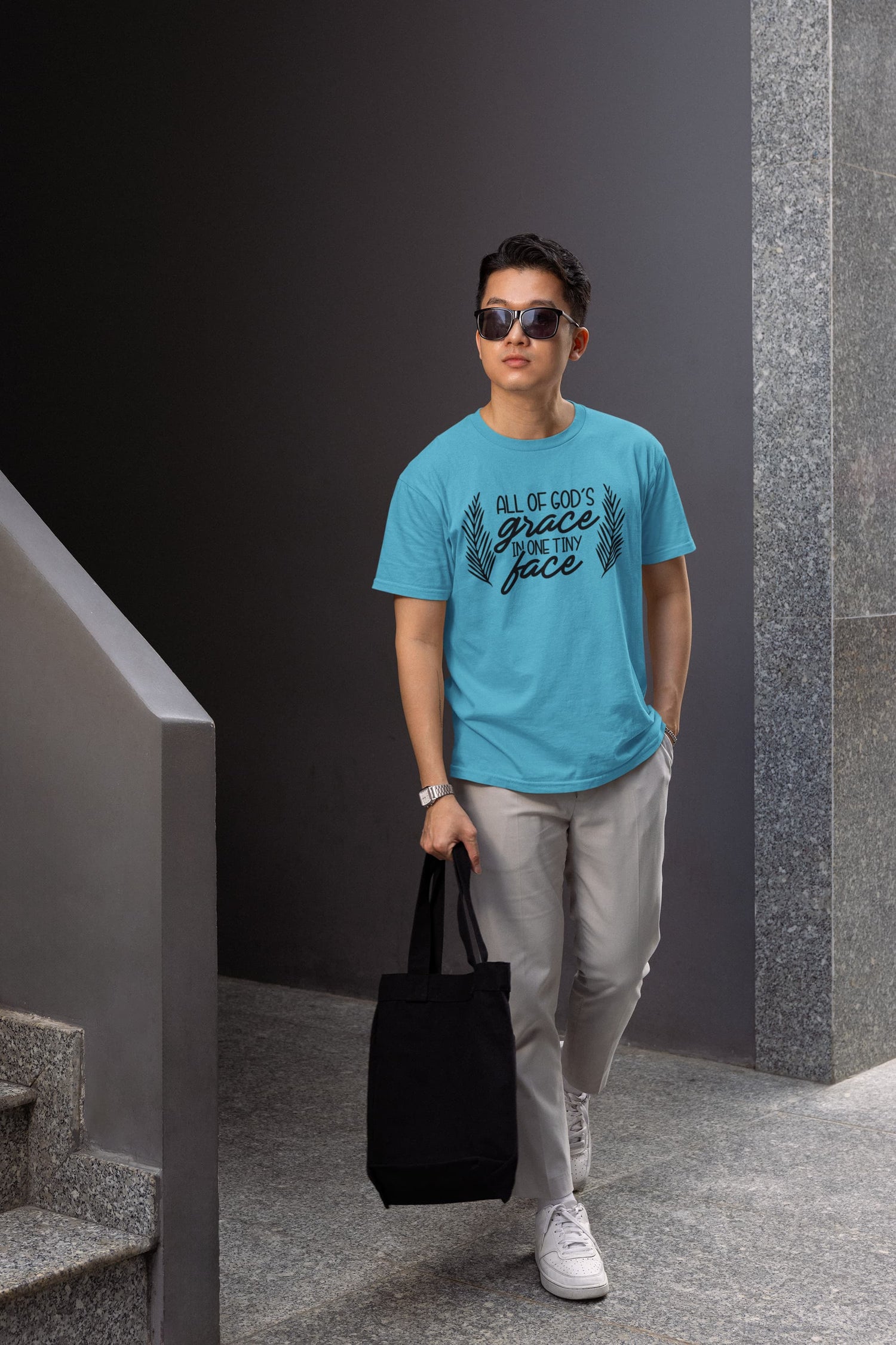 Man wearing a blue t-shirt with text, holding a black bag, standing against a dark wall.