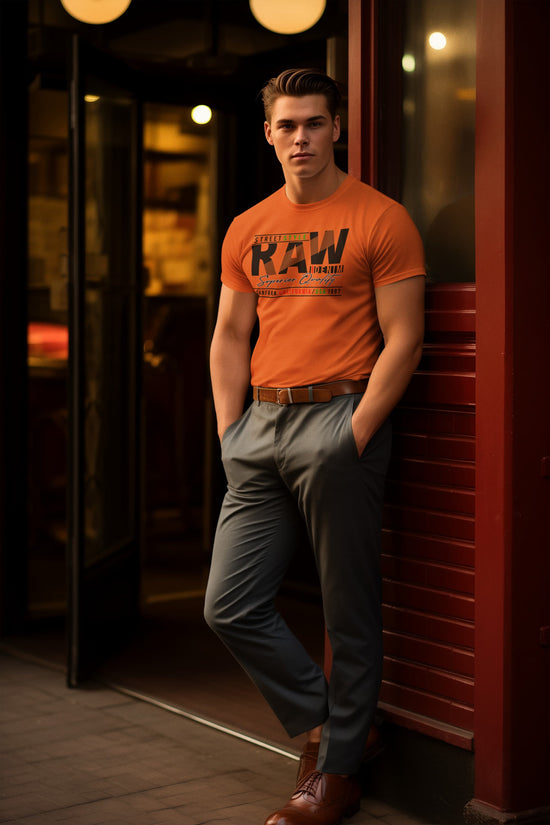 Man wearing an orange t-shirt with text and gray pants leaning against a red wall.