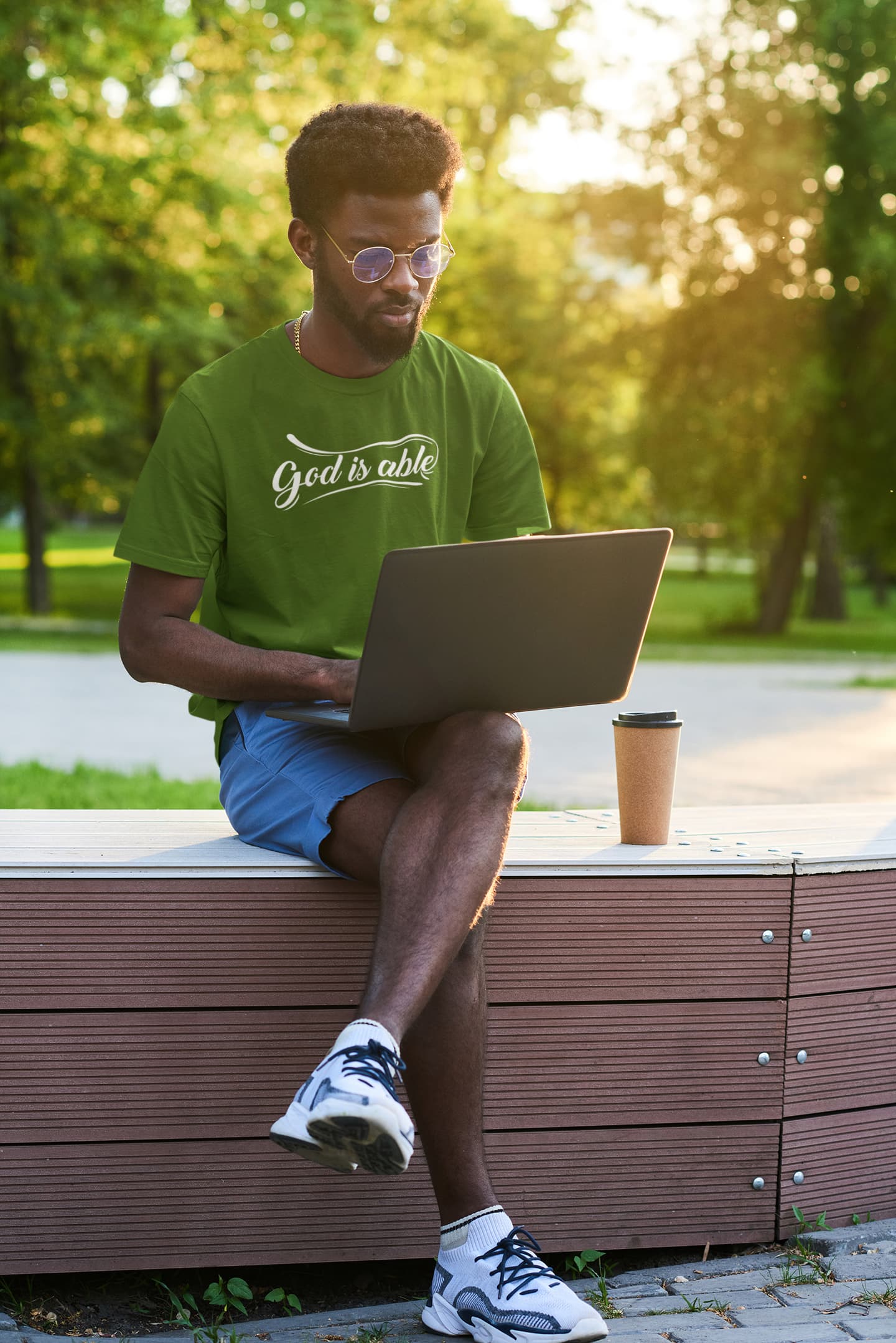 Man sitting on a bench outdoors using a laptop with a green shirt and sunglasses.