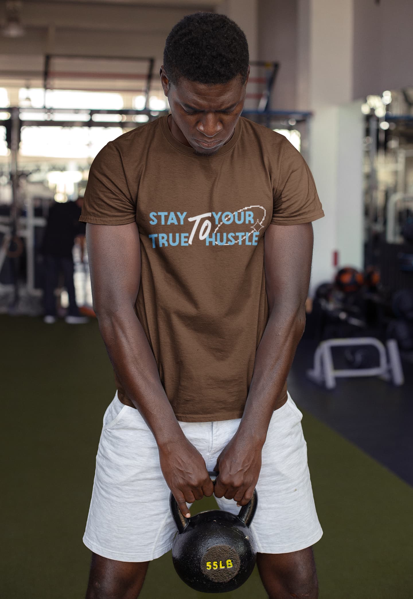 Person lifting a kettlebell in a gym wearing a brown t-shirt with text.