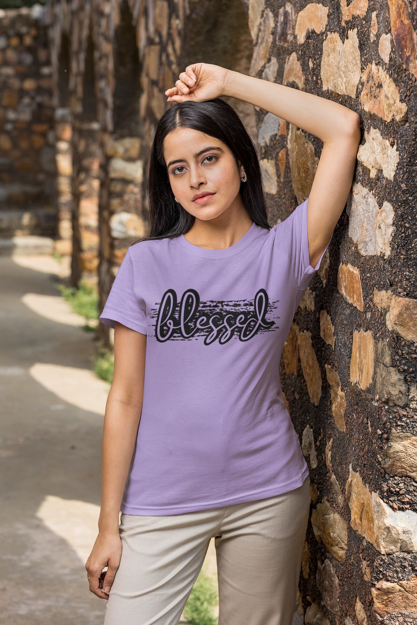 Woman wearing a purple t-shirt with 'blessed' text, standing against a stone wall.