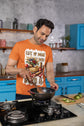 Man cooking in a kitchen wearing an orange t-shirt with a graphic design.