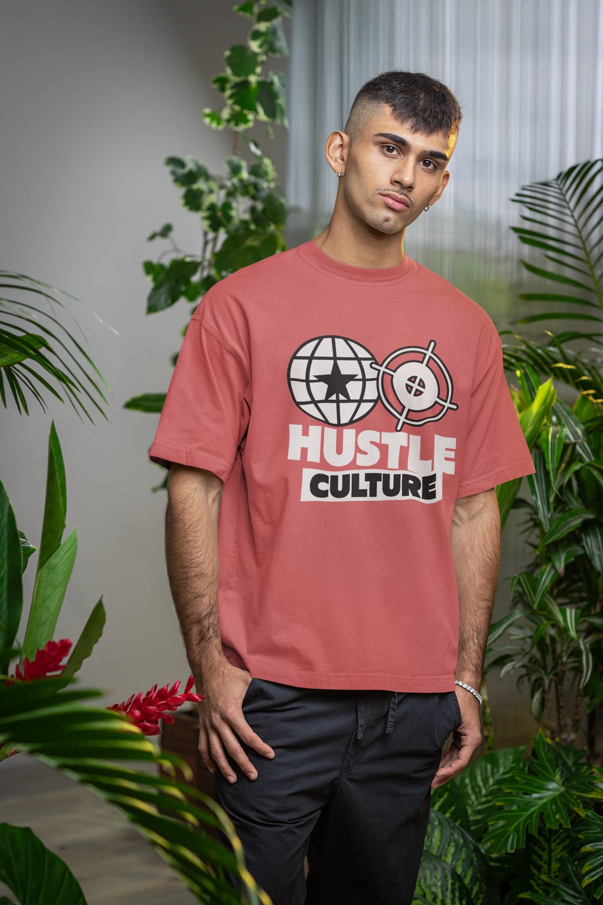 Man wearing a red t-shirt with 'Hustle Culture' graphic design, surrounded by green plants.