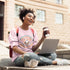 Woman sitting on steps with a laptop and coffee cup, wearing a pink shirt and jeans.