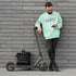 Man in a light green sweatshirt standing next to a scooter against a gray brick wall.