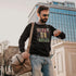 Man wearing a black sweatshirt with text, holding a beige bag, standing in an urban setting.