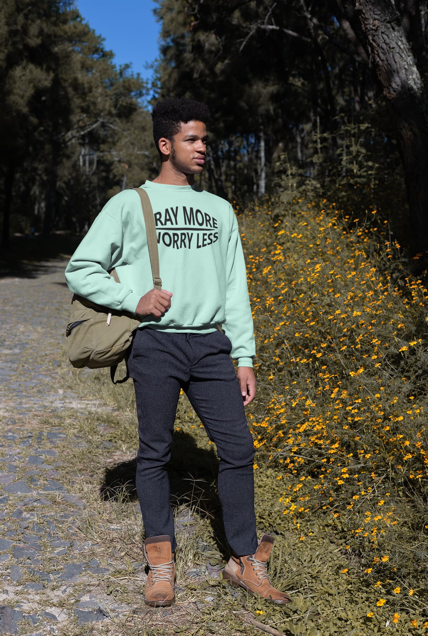 Man standing outdoors with a green sweatshirt and bag, surrounded by trees and yellow flowers.