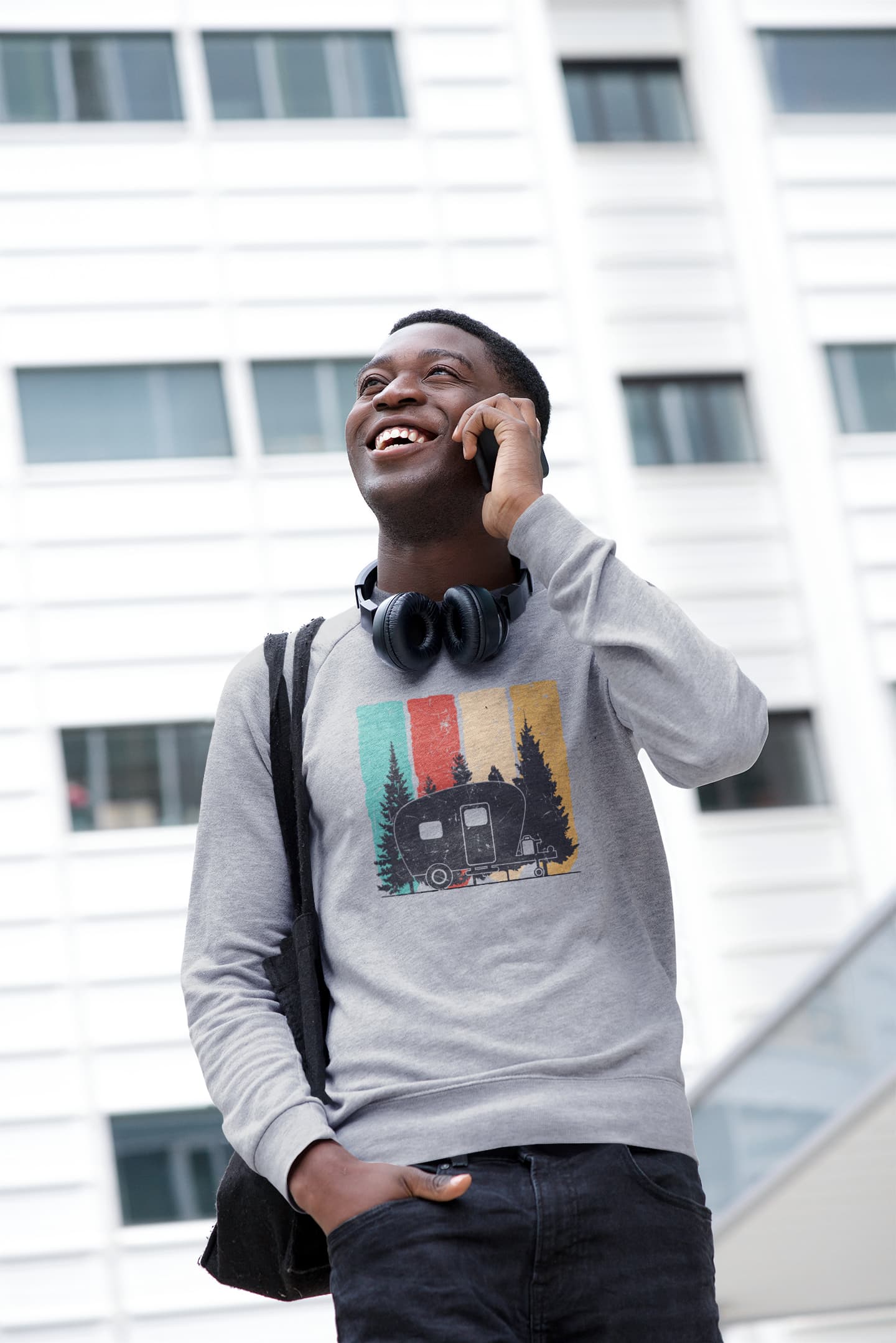 Man talking on phone with a backpack and headphones, wearing a graphic sweatshirt, in front of a building.