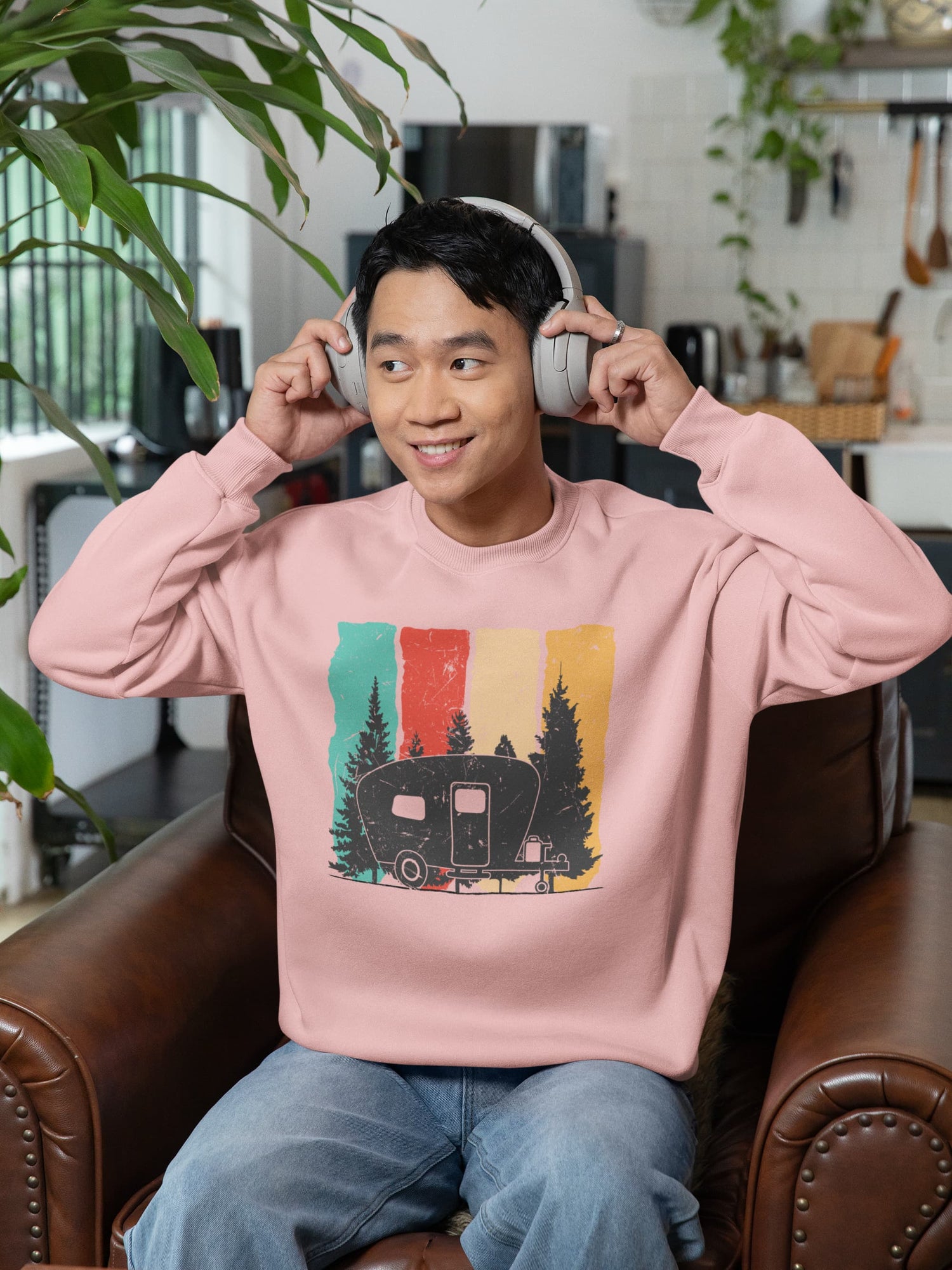 Person wearing a pink sweatshirt with a graphic design, sitting on a couch in a room with plants and kitchen items.