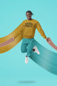 Person wearing a mustard yellow sweatshirt with text, teal pants, and white shoes on a teal background