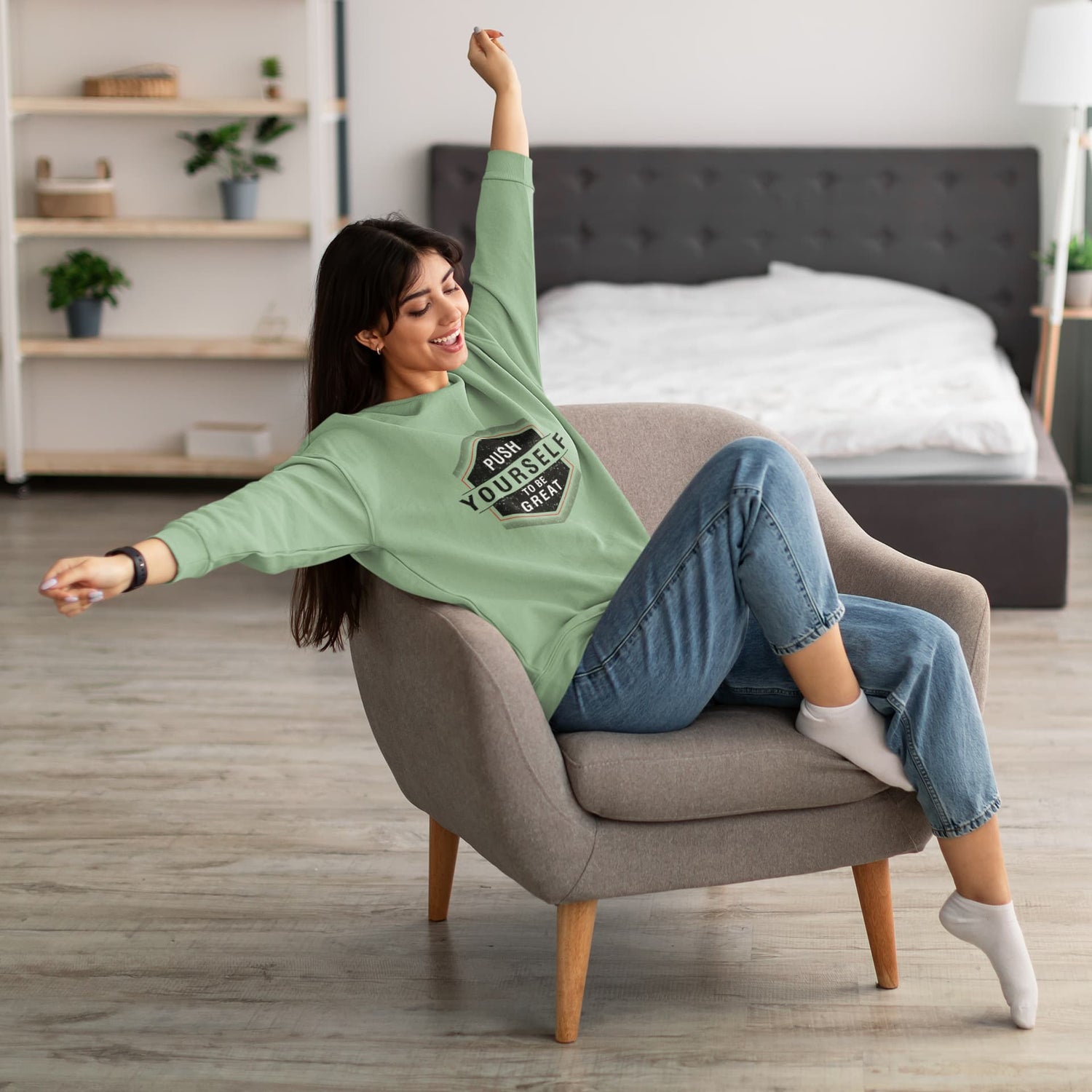 Woman sitting in a chair wearing a green sweatshirt and blue jeans in a bedroom.