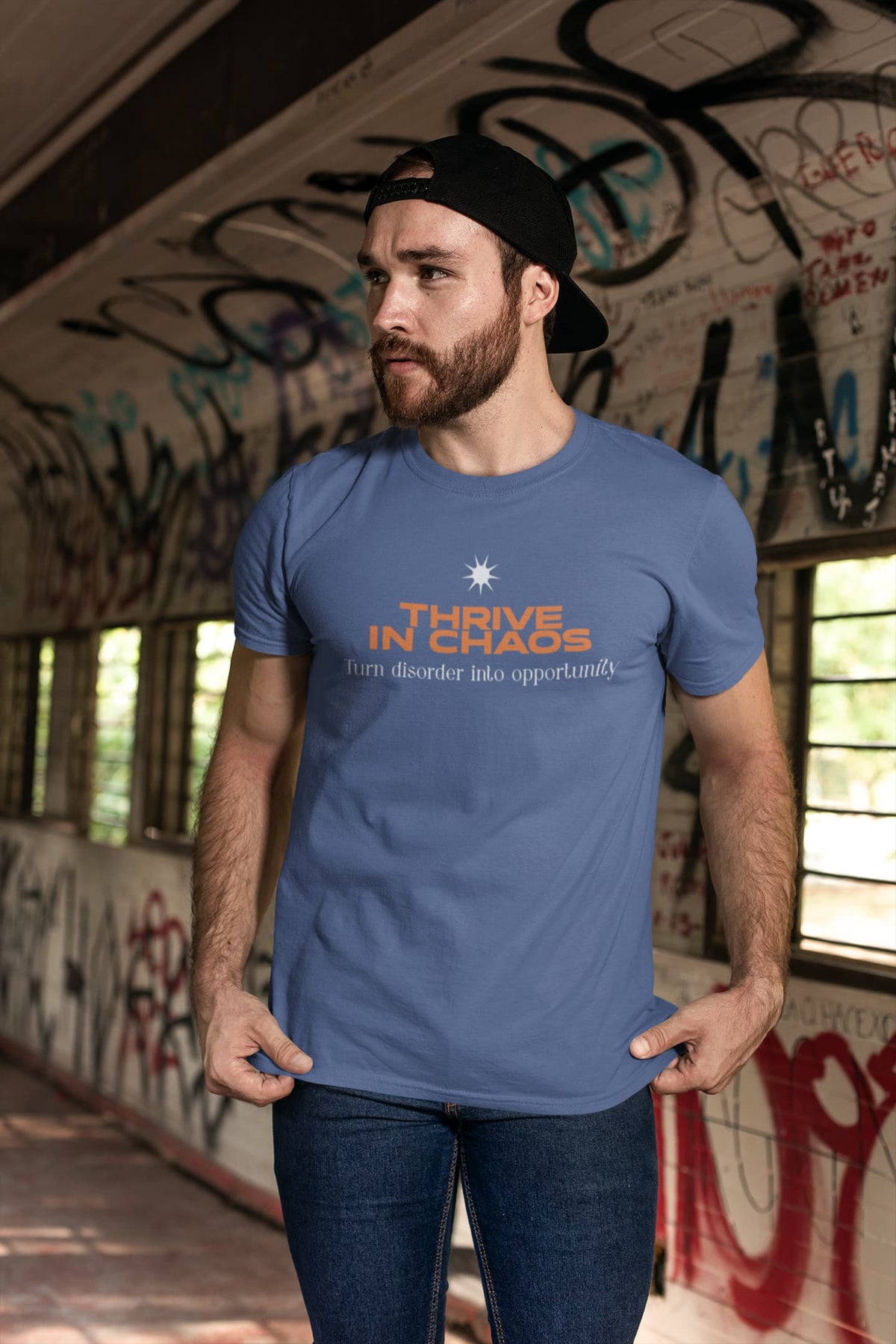 Man wearing a blue t-shirt with 'Thrive in Chaos' text in an urban setting with graffiti.