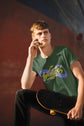 Person wearing a green t-shirt with a logo, holding a skateboard, against a blurred background