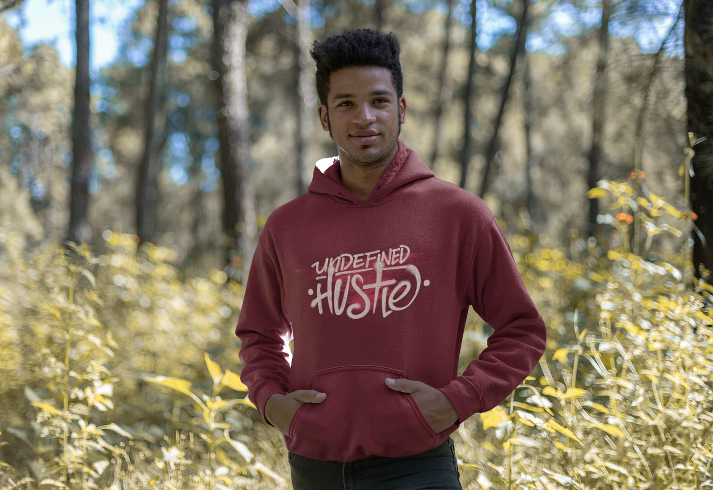 Person wearing a maroon hoodie with 'UNDEFEATED HUSTLE' text in a forest setting