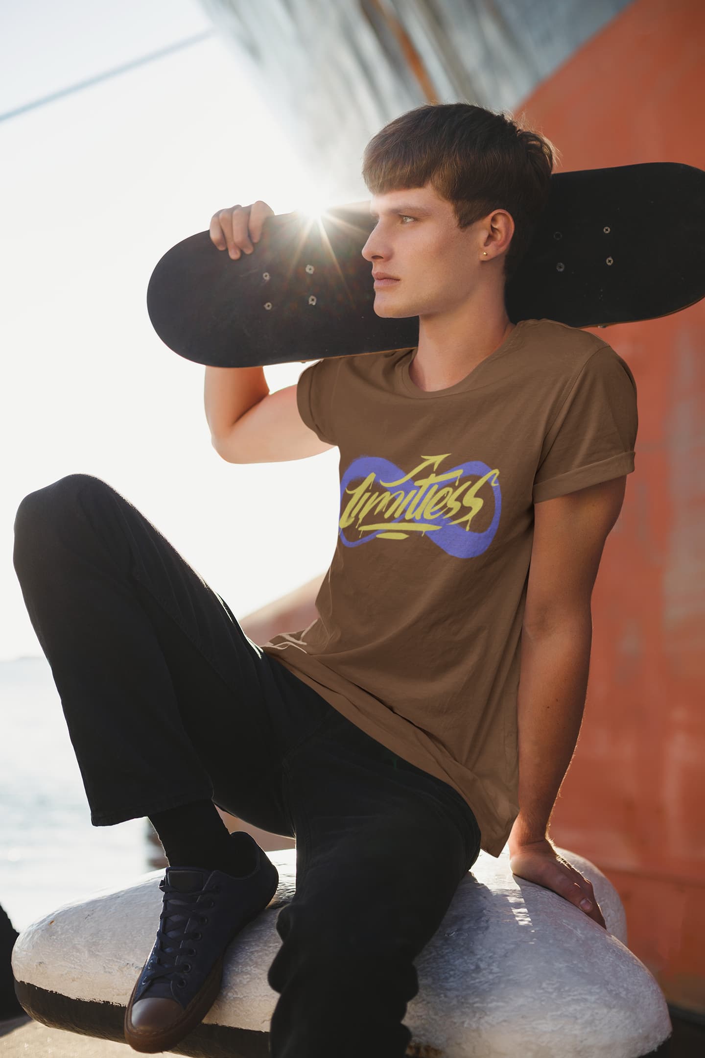 Person wearing a brown t-shirt with a logo, holding a skateboard, sitting on a rock by the water.