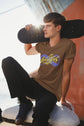 Person wearing a brown t-shirt with a logo, holding a skateboard, sitting on a rock by the water.