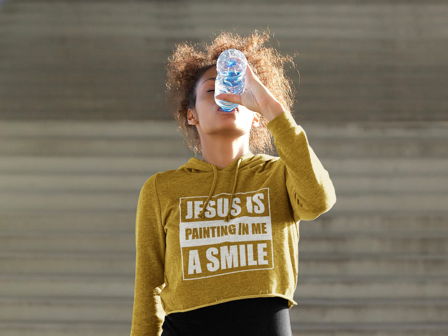 Person wearing a mustard yellow hoodie with text, drinking from a water bottle against a plain background