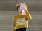 Person wearing a mustard yellow hoodie with text, drinking from a water bottle against a plain background
