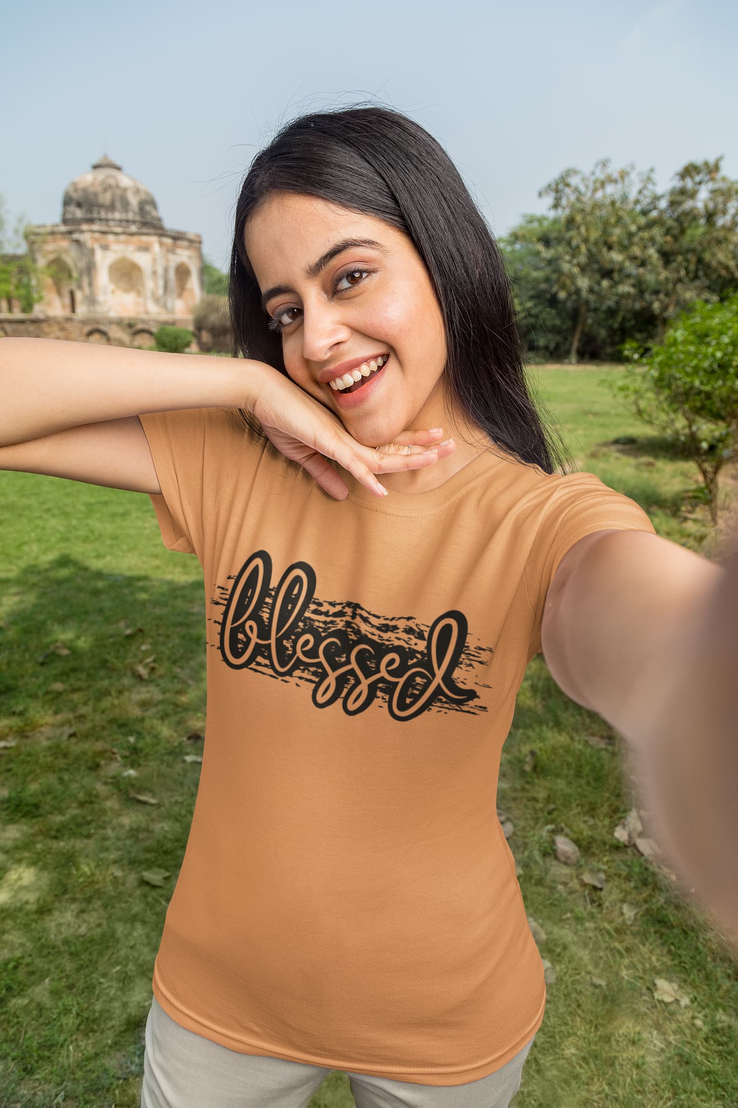 Woman wearing a brown t-shirt with 'blessed' text, standing outdoors with greenery and a building in the background.