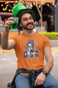 Man wearing an orange t-shirt with a graphic design, holding a glass of green liquid, and wearing a green hat.